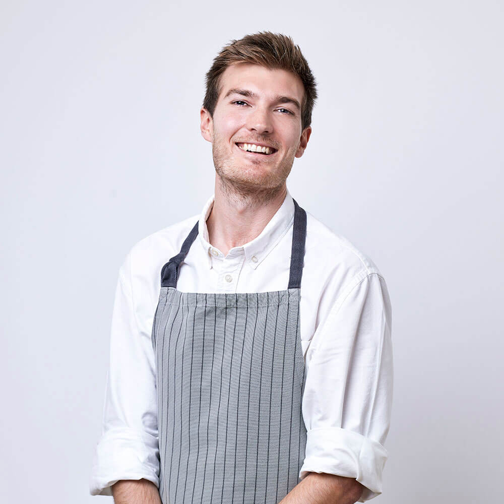 Photograph of a kitchen assistant wearing a white shirt and an apron, looking at the camera and smiling.