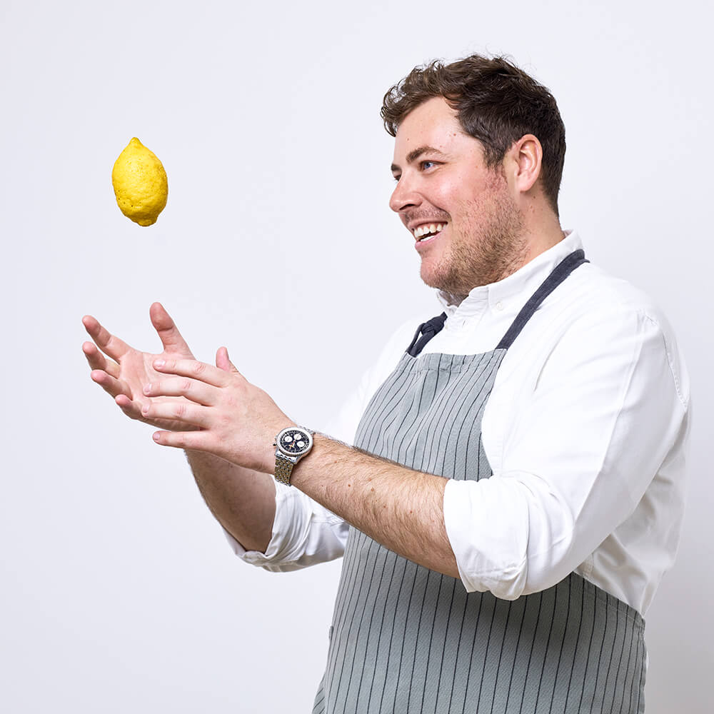 Photograph of a chef wearing a white shirt and grey and black striped apron. He is throwing a lemon in the air and smiling.