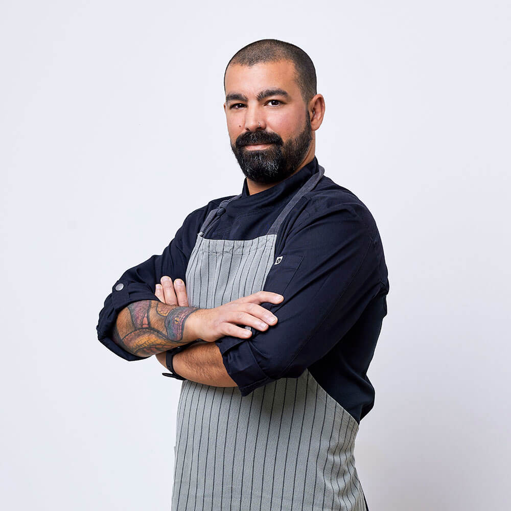 Photograph of a chef wearing a black shirt and a grey and black striped apron. He is crossing his arms and looking at the camera.