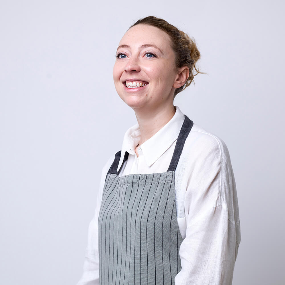 Photograph of a kitchen assistant wearing a white shirt and an apron, looking at the camera and smiling.