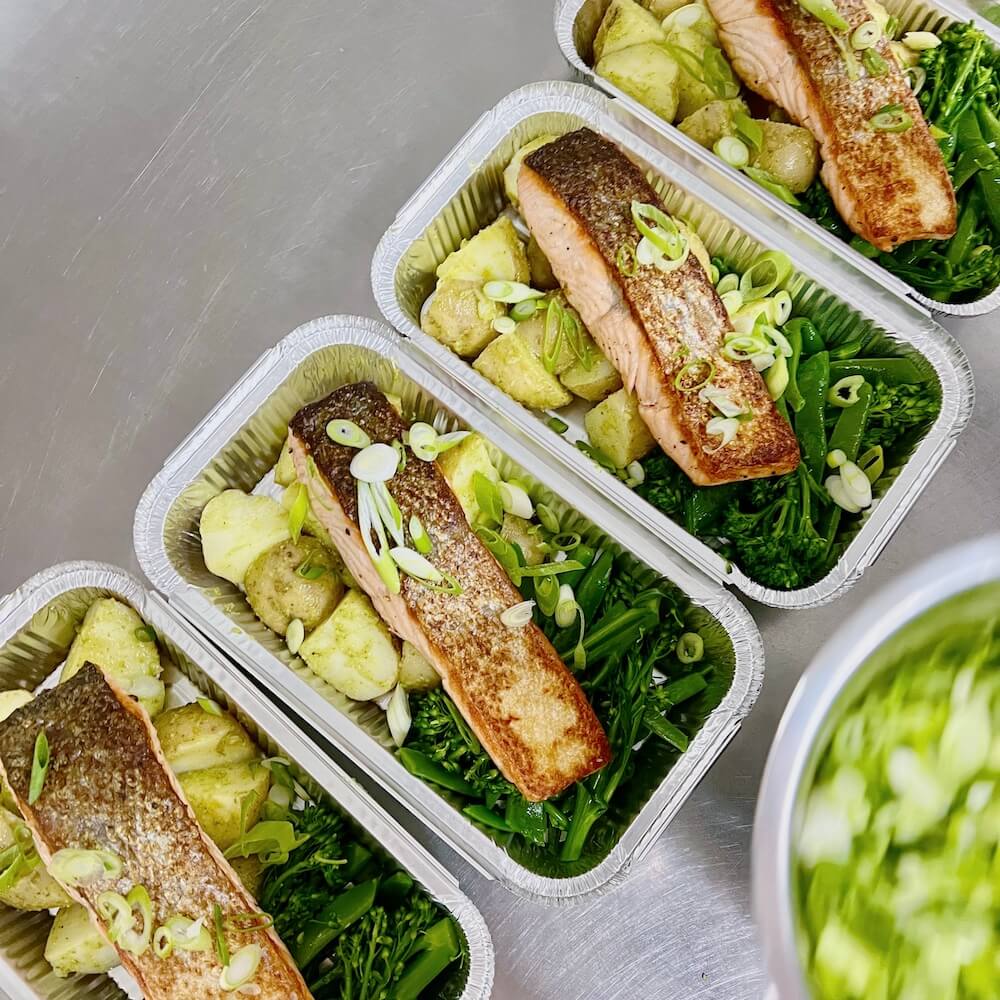 Photograph from above of several meal prep food takeaway trays containing salmon fillets with spring onions, roast potatoes and sautéed greens