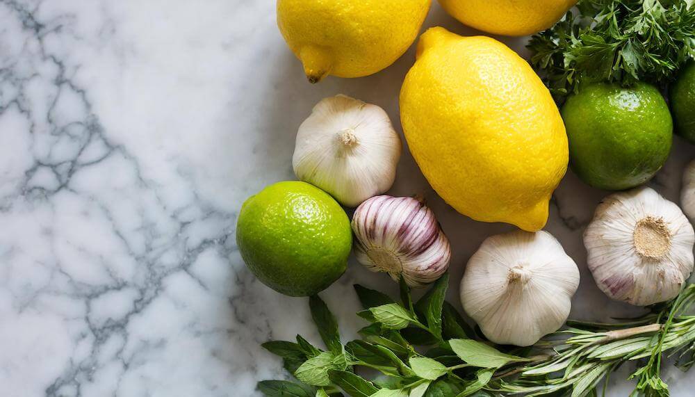 Photograph from above of a white marble kitchen counter with lemons, limes, bulbs of garlic and sprigs of mint scattered on top