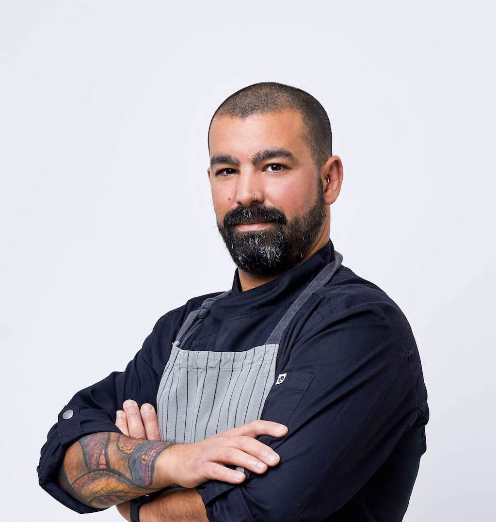 Photograph of a chef stood with his arms crossed in a black chefs shirt and grey and black striped apron