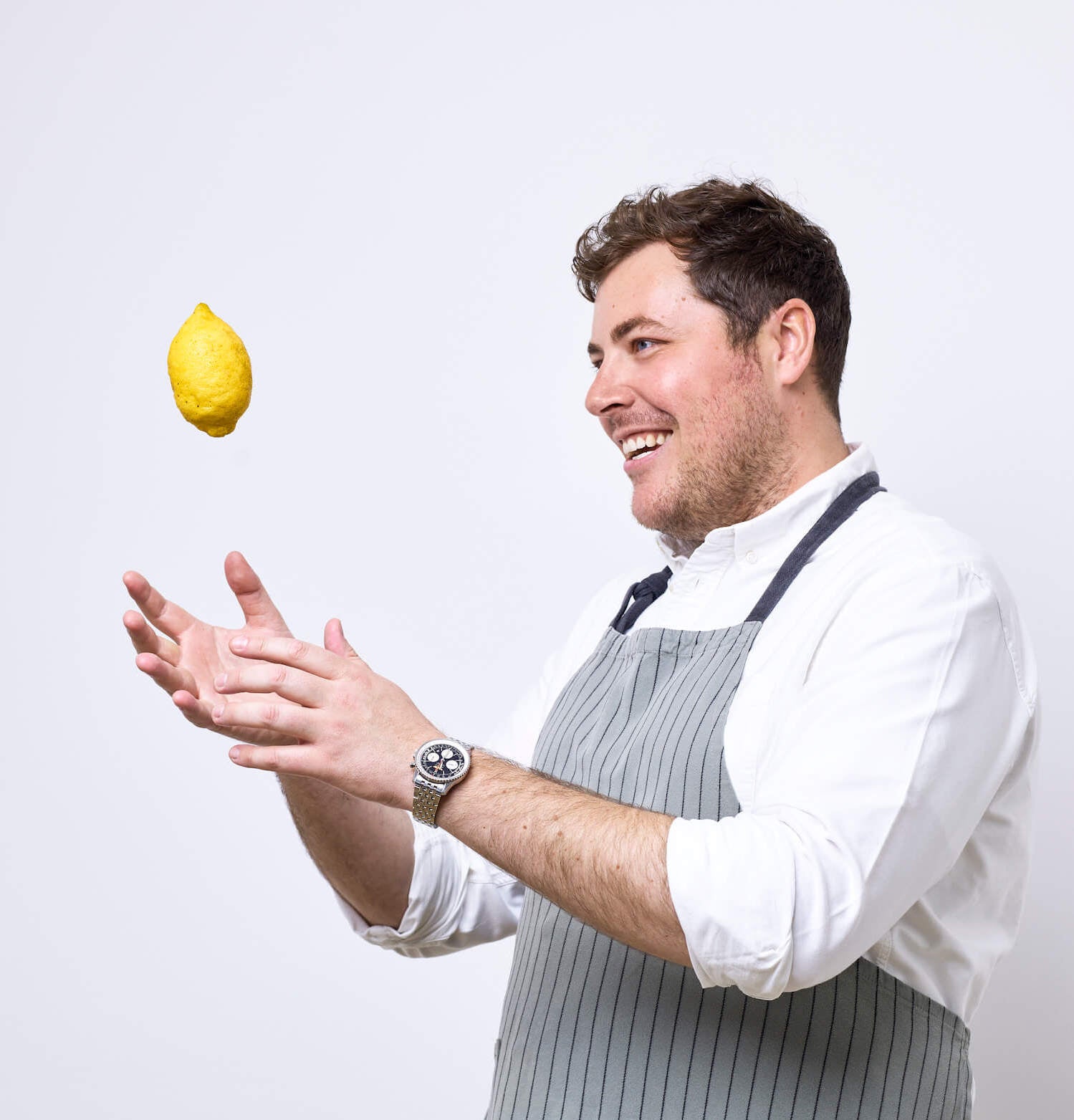 Photograph of a chef in a white shirt and grey and black striped apron. The chef is throwing a lemon in to the air and smiling