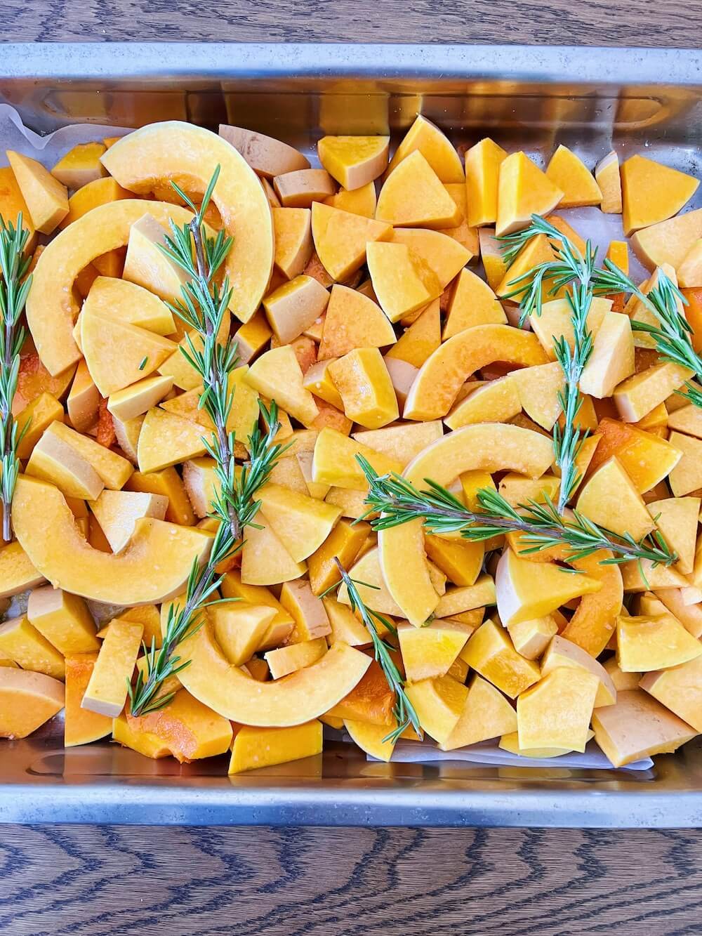 Photograph of chopped up butternut squash in an oven tray with sprigs of rosemary on top, prepared for cooking