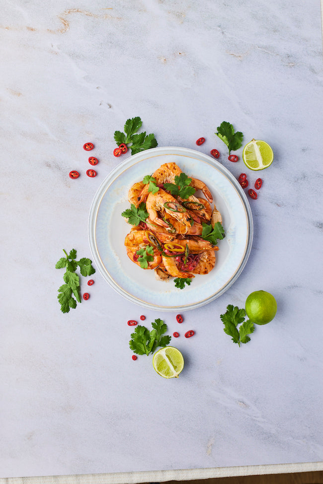 Photograph from above of a plate of chilli and garlic prawns. The plate is on a white marble surface and is surrounded by limes (cut in half or whole), coriander leaves and slices of red chilli